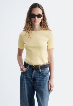 Femme aux longs cheveux ondulés portant des lunettes de soleil en écaille, un haut jaune à manches courtes, un jean bleu avec une ceinture marron et des bijoux en argent, debout devant un fond blanc.