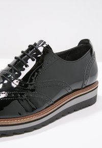 Chaussure en cuir verni noir avec des détails de brogue, bout rond et laçage à l'avant. Présente une semelle plateforme empilée avec des accents marron et gris.