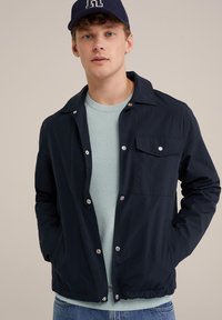 Mornarska lahka jakna s zaklopnimi gumbi, žepom na prsih in elastičnim robom, oblečena čez svetlo modro teksturirano sveternico in kavbojke.