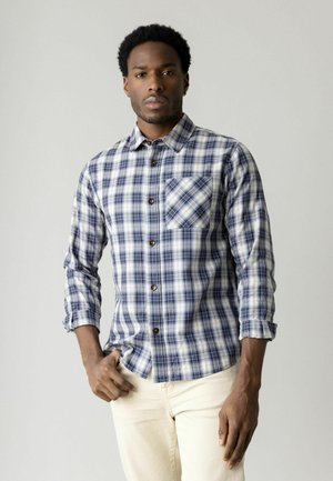 Mann trägt ein blau-weiß kariertes Hemd mit langen Ärmeln und hellbeige Hose, steht mit einer Hand in der Tasche vor einem schlichten Hintergrund.