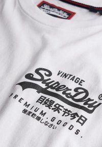 Camiseta de algodón blanca con un estampado gráfico negro prominente con la palabra "SuperDry" y texto adicional en japonés. Diseño de cuello redondo con etiqueta visible.