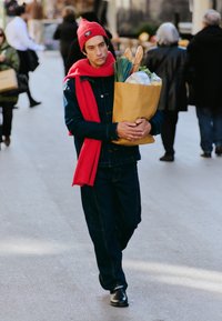 Veste en denim et pantalon foncé, bonnet rouge et écharpe ; portant un sac en papier brun rempli de courses, comprenant des baguettes et des légumes.
