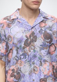 Homme portant une chemise à manches courtes boutonnée avec un motif floral multicolore dans les tons violet, rose, blanc et marron.