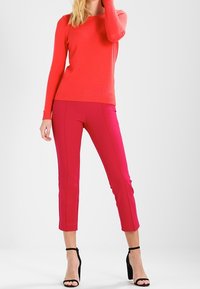 Rode gebreide trui en roze op maat gemaakte broek, met een slim fit en cropped lengte. Zwarte hakken maken de outfit compleet.