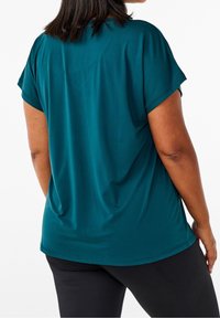 Teal kortærmet top lavet af glat stof, med en afslappet pasform og rundet kant, parret med sorte bukser, set fra bagsiden.