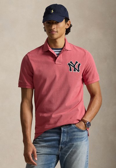 Homme portant une casquette bleu marine, un polo rose saumon avec le logo des New York Yankees, un maillot rayé en dessous, un jean bleu et une montre-bracelet, debout avec une main dans la poche.