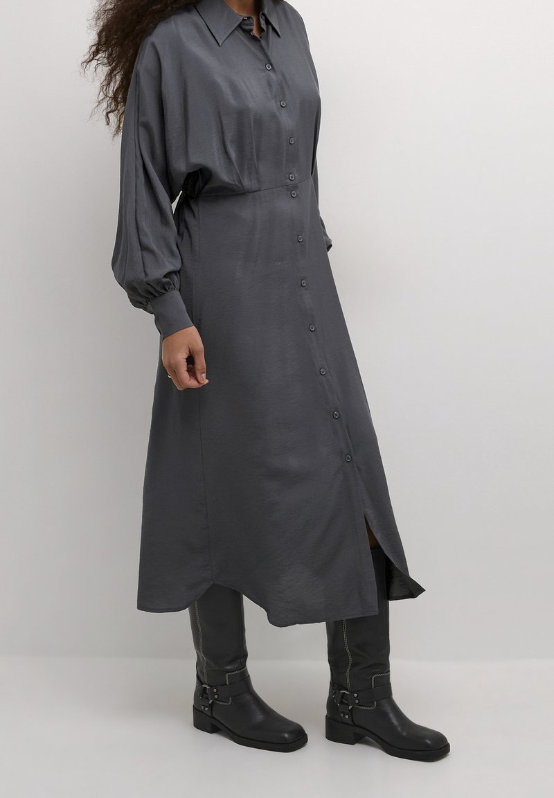Femme portant une robe midi gris foncé à manches longues et boutonnée, associée à des bottes noires en cuir jusqu'aux genoux, debout devant un fond uni.