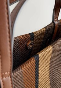 Tkaná nákupná taška v prírodných tónoch hnedej, béžovej a čiernej s koženými doplnkami. Má dekoratívne zapínanie na gombík a textúrovaný vzor.