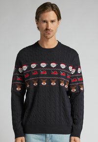 Granatowy sweter z dzianiny z świątecznym wzorem przedstawiającym twarze Świętego Mikołaja, czerwone ciężarówki i renifery. Wzór warkocza na ciele i rękawach.