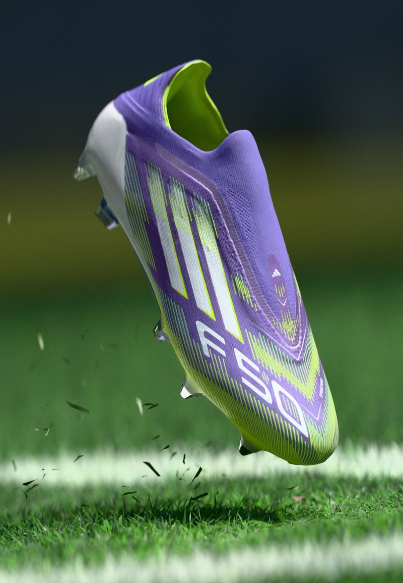 Scarpa da calcio viola e verde con un design aerodinamico, parte superiore testurizzata e tacchetti visibili, sospesa sopra l'erba con frammenti di erba volante.