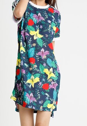Person iført en løs mørkeblå kjole med farverige blomsterprint, herunder gule, lilla og røde blomster samt grønne blade, stående mod en ensfarvet baggrund.