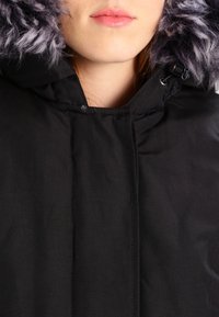 Chaqueta negra con capucha que cuenta con un borde de piel sintética texturizada, con cierre de cremallera y cordones ajustables para un ajuste ceñido.