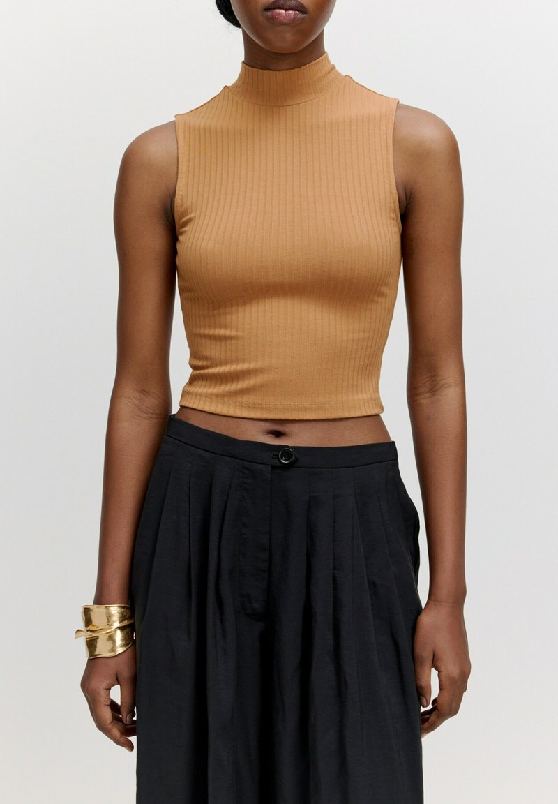 Brauner gerippter ärmelloser Crop-Top mit hohem Halsausschnitt, kombiniert mit einem schwarzen plissierten Rock. Besitzt einen einfachen Knopfverschluss an der Taille.