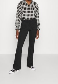 Černé rozšířené kalhoty s hladkým texturem, zkombinované s černobílou kostkovanou blůzou se volánovými manžetami. Dokončeno s černými teniskami.