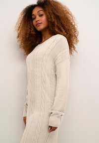 Rochie tricotată de culoare bej deschis, cu modele de tricotaj cu cablu, mâneci lungi cu tivuri ribbate și o croială relaxată.