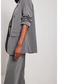 Veste de costume grise à fines rayures avec une coupe décontractée. Elle présente de grands revers, deux boutons et des manches retroussées. Tissu lisse avec une texture subtile.