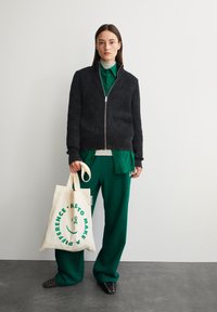 Schwarzer gerippter Reißverschluss-Pullover, darunter ein grünes Hemd mit Kragen, grüne weite Hose und eine cremefarbene Tragetasche mit grünem Text und Smiley-Design.