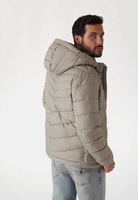 Jachetă puffer de un gri deschis, cu un model matlasat, având glugă și manșete elastice. Purtați peste blugi de un bleumarin deschis.