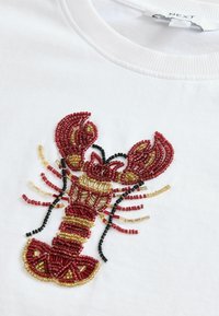 Tricou alb cu un design de homar din mărgele roșii și aurii, cu accente negre, evidențiind lucrări de mărgele complexe și o aparență texturată.