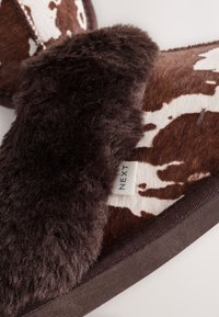 Brązowo-białe kapcie w wzory, z miękką podszewką z sztucznego futra i ciemnobrązowym obszyciem. Na boku widoczna metka z napisem „NEXT”.
