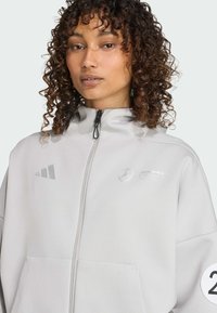 Hellgraue Zip-Jacke mit hohem Kragen, ausgestattet mit Adidas- und Mercedes-Logos, seitlichen Taschen und einem runden Aufnäher mit der Nummer 23.