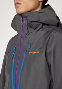 Mężczyzna w ciemnoszarej kurtce Patagonia z wysokim kołnierzem, fioletowym zamkiem z przodu, niebieskimi zamkami kieszeni i pomarańczowym logo na piersi.