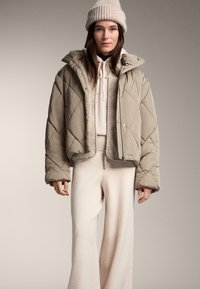 Béžová prešívaná bunda s kapucňou, ktorá má mäkkú podšívku. Kombinovaná so svetlo béžovou mikinou a širokými nohavicami, doplnená pletenou čiapkou.