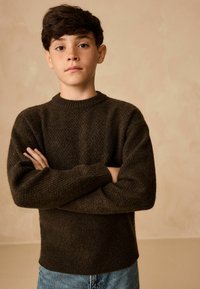 Brązowy sweter dziany z teksturowanym splotem i ściągaczami na rękawach, połączony z niebieskimi dżinsami. Model ma skrzyżowane ręce na neutralnym tle.