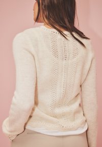 Suéter de punto beige claro con un delicado diseño de encaje en la parte posterior, puños acanalados y una textura suave, superpuesto a una blusa blanca.