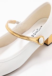 Ballerina bianca in pelle con una fascia dorata decorata da un bottone rotondo e un piccolo tacco quadrato. Texture liscia e design minimalista.