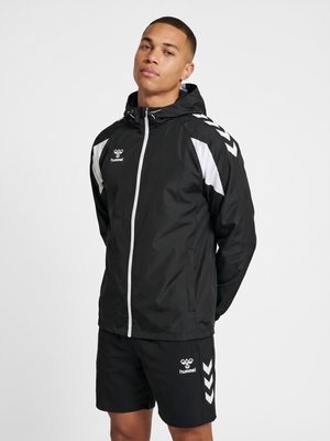 Man draagt een zwart-wit Hummel sportjack met capuchon en bijpassende zwarte shorts tegen een effen lichte achtergrond.