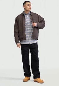 Giacca marrone, camicia a righe, pantaloni neri e stivali color sabbia. La giacca presenta una tasca frontale e una vestibilità ampia; la camicia ha righe blu e bianche.