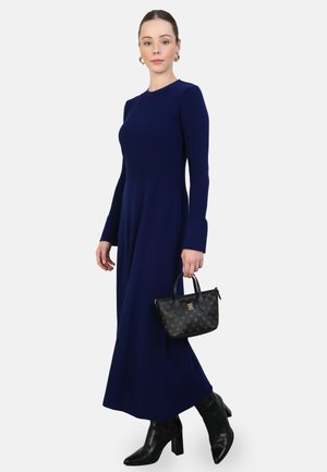 Knit navy dress with long sleeves, textured bodice, and smooth skirt. Model holding a small black handbag with subtle pattern. Black heeled boots.