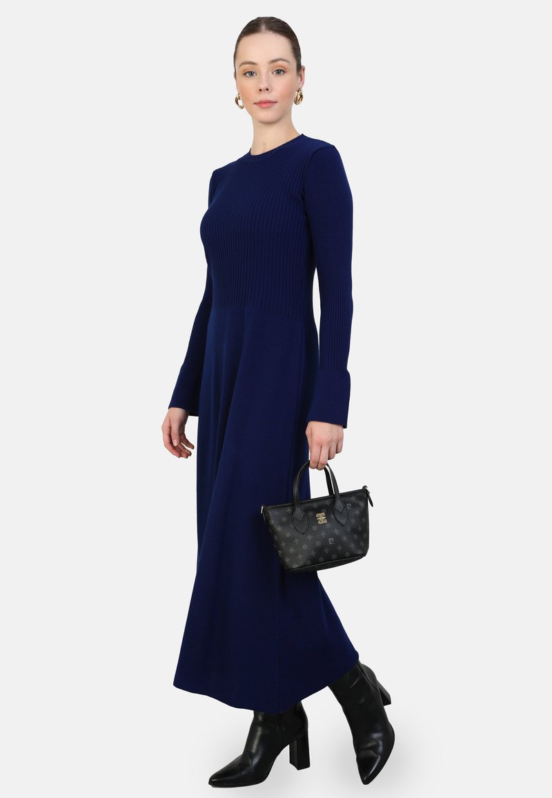 Abito lavorato a maglia blu navale con maniche lunghe, corpino testurizzato e gonna liscia. Modella che tiene una piccola borsa nera con un motivo sottile. Stivali neri con tacco.