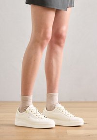 Vita lädersneakers med gulddetaljer, rund tå och tjocka sulor. Bärs med beige ribbade strumpor, placerade mot ett ljust trägolv.