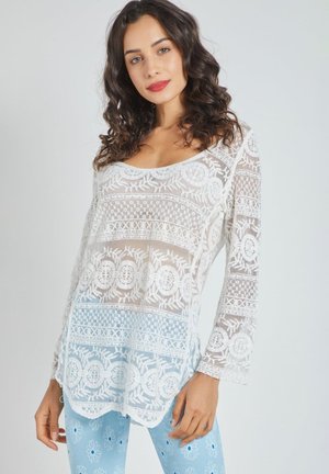 Blusa de encaje blanco con patrones florales, mangas largas y escote en forma de u, llevada sobre leggings de estampado azul claro. Tejido texturizado y transparente.