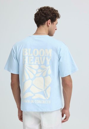 Mies, jolla on vaaleansininen t-paita, jossa on kermaan vivahtava teksti "BLOOM HEAVY" ja abstrakteja muotoja, yhdistettynä valkoisiin housuihin, kasvoin poispäin kamerasta.