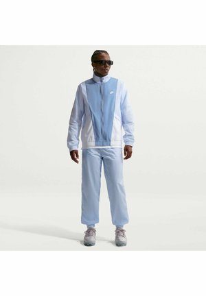 Mann mit geflochtenen Haaren, der dunkle Sonnenbrillen und hellblaue Nike-Sportbekleidung trägt, steht mit locker hängenden Händen an den Seiten vor weißem Hintergrund.