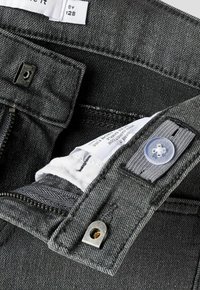 Gros plan sur la ceinture d'un jean gris foncé montrant un bouton pression, un crochet en métal, une fermeture éclair et une étiquette d'entretien visible à l'intérieur.