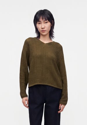 Jeune femme aux cheveux courts noirs portant un pull texturé vert olive à manches longues et un pantalon ample foncé sur fond blanc.