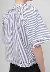 Femme portant un chemisier à manches courtes rayé verticalement bleu et blanc avec des découpes décoratives et une bordure à pompons, associé à un pantalon noir.