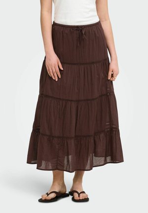 Femme portant une jupe longue marron à volants avec des détails en dentelle, associée à des tongs noires et un haut blanc sans manches.
