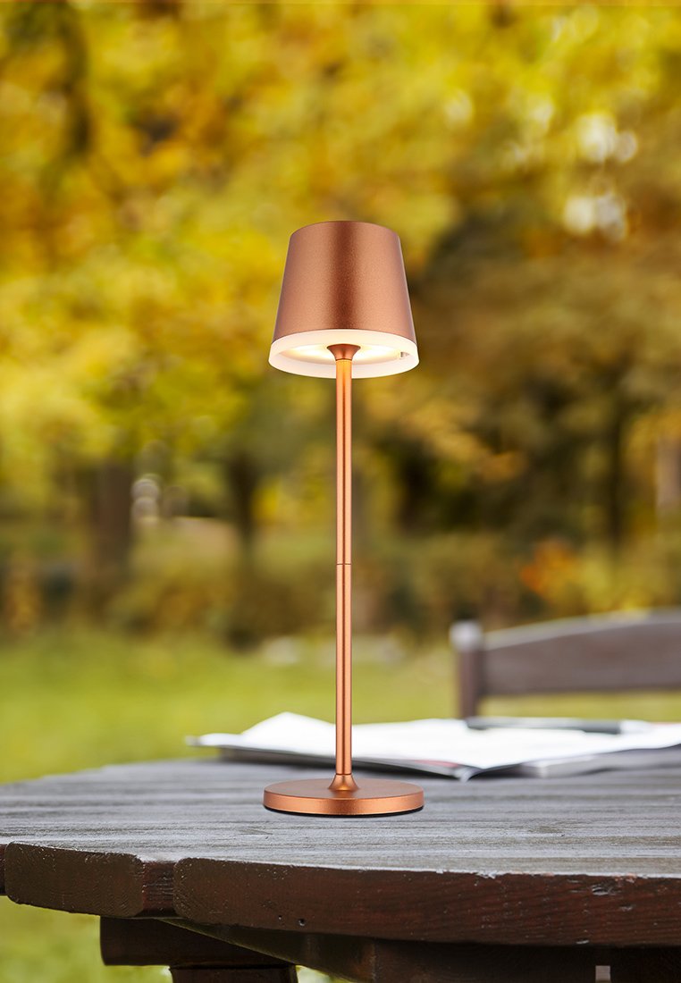 Kupferne Tischlampe mit einem zylindrischen Schirm, schlankem vertikalem Ständer und runder Basis. Glatte Metalloberfläche; vor einem verschwommenen, äußeren Hintergrund.