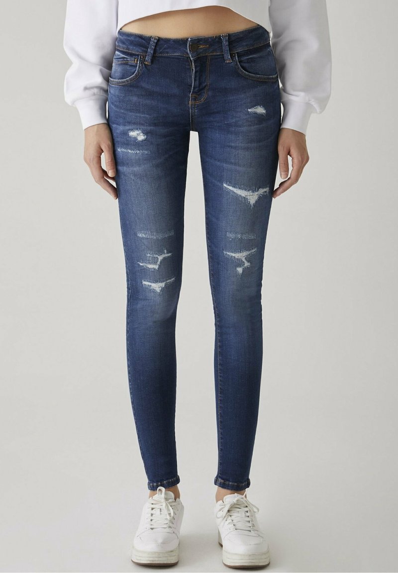 Jean skinny en denim bleu foncé avec des détails usés, comprenant des déchirures sur les cuisses et une taille mi-haute. Porté avec des baskets blanches.