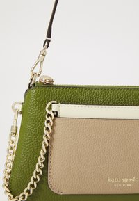 Borsa verde in pelle con tasca frontale color cuoio, hardware dorato e tracolla a catena. Presenta una finitura testurizzata e un dettaglio del logo incassato.