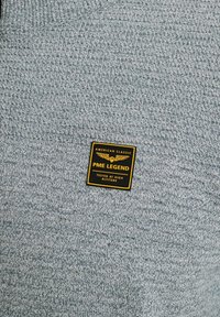 Grijze gebreide trui met een textuurpatroon en een zwart-gouden label waarop "PME LEGEND" staat, met "AMERICAN CLASSIC" en "TESTED AT HIGH ALTITUDE."