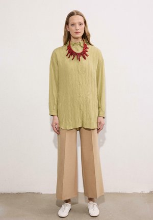 Woman stands wearing a loose light green buttoned shirt, beige wide-leg pants, white shoes, and a chunky red necklace against a plain wall.