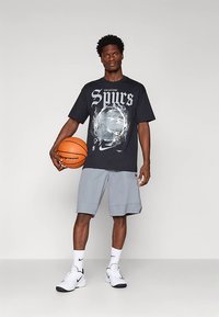 Vīrietis stāv, labajā rokā turot basketbola bumbu, ģērbies melnā San Antonio Spurs T-kreklā, pelēkās šortos, baltās Nike zeķēs un baltos sporta apavos.