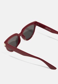 Gafas de sol de gato rojo con lentes negros, marco de plástico brillante y sutiles acentos dorados en las bisagras. Diseño ligero y elegante.