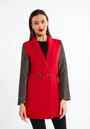 Femeie purtând un blazer roșu fără mâneci cu mâneci lungi negre și aurii cu model, top negru și cercei triunghiulari, stând în fața unui fundal alb.